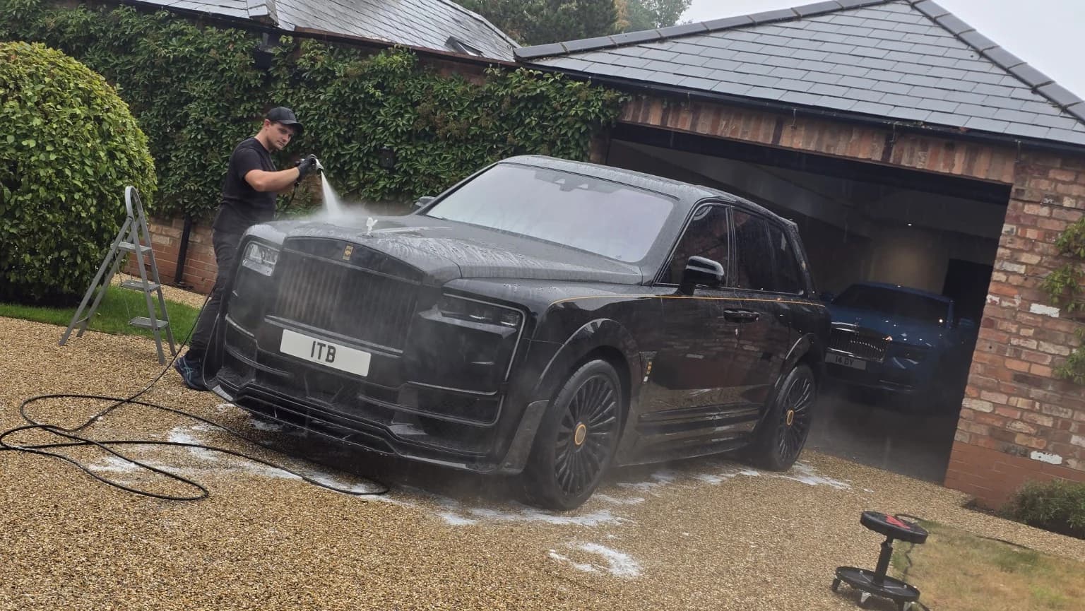 A detailer rinsing a black Rolls-Royce on a private driveway